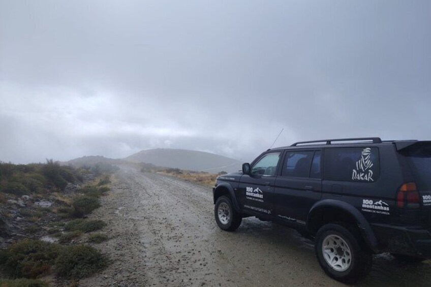 A black SUV on a gravel road with 'Dakar' branding, surrounded by sparse vegetation and hills in the background. 4