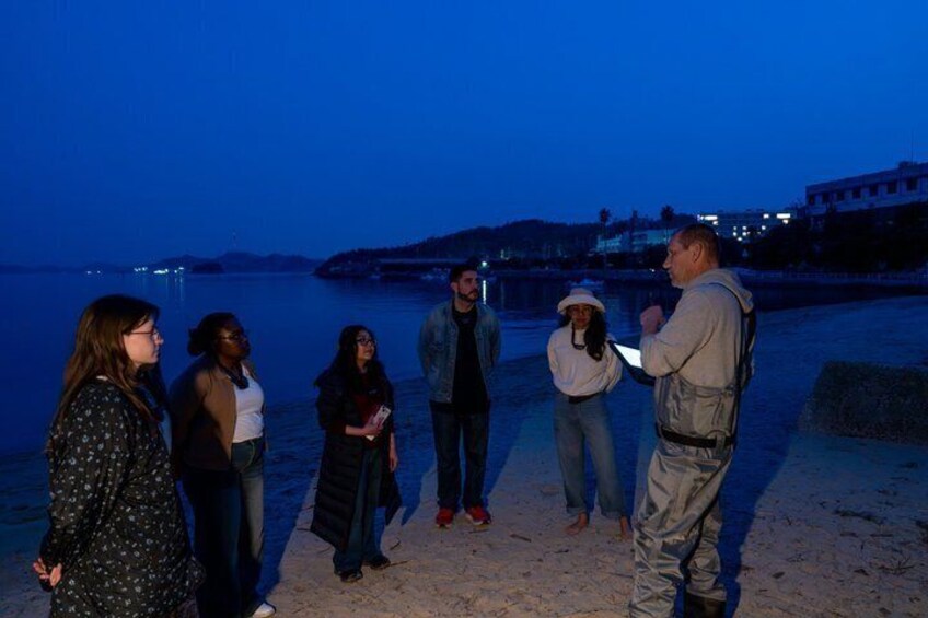 A group of people standing on a beach at dusk with a body of water and buildings in the background. 3