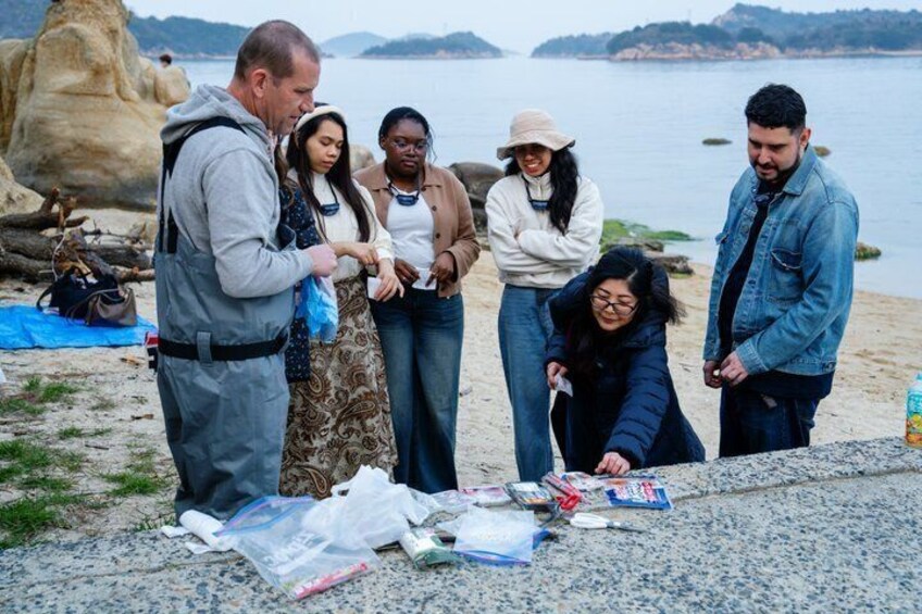 A group of people standing on a rocky shore by the sea, some holding items. 5