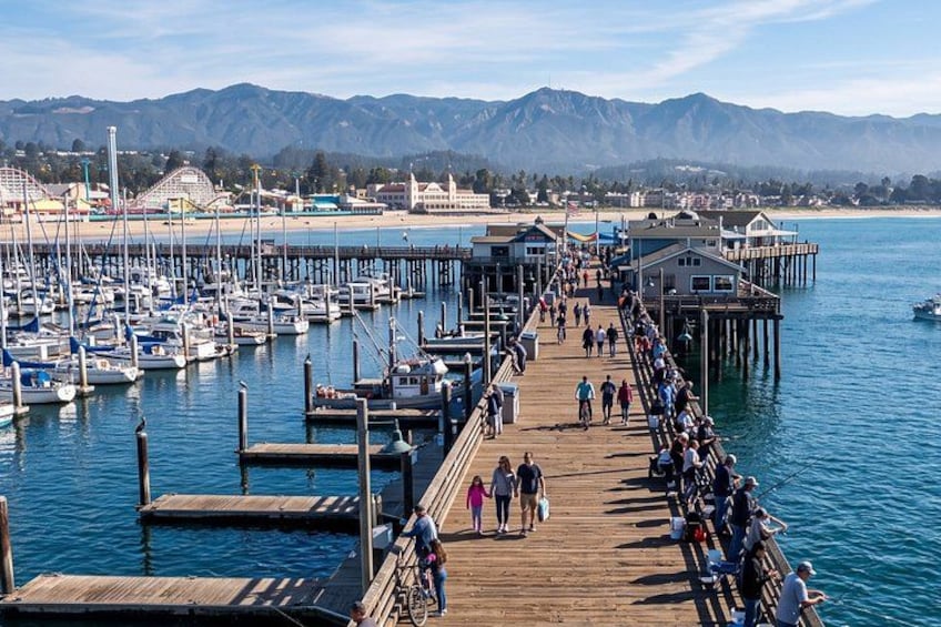 A pier with people walking and cycling, boats docked, and mountains in the background. 1