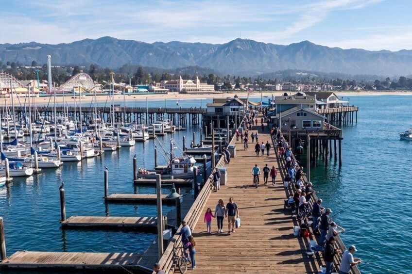A pier with people walking and cycling, a marina with boats, and mountains in the background. 1