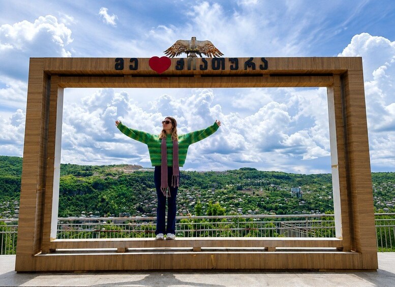 A person standing in a wooden frame with arms outstretched, overlooking a green landscape with hills and a clear sky. 2