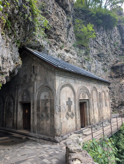A stone building with cross motifs and a tiled roof, set against a rocky cliff. 3