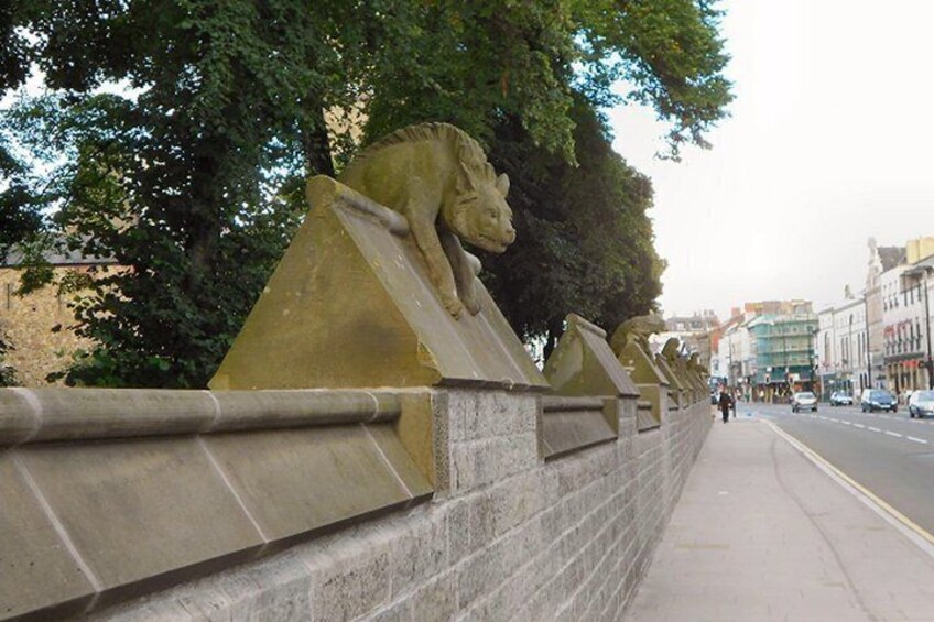 A stone wall with lion statues, a sidewalk, and a street with cars and buildings in the background. 5