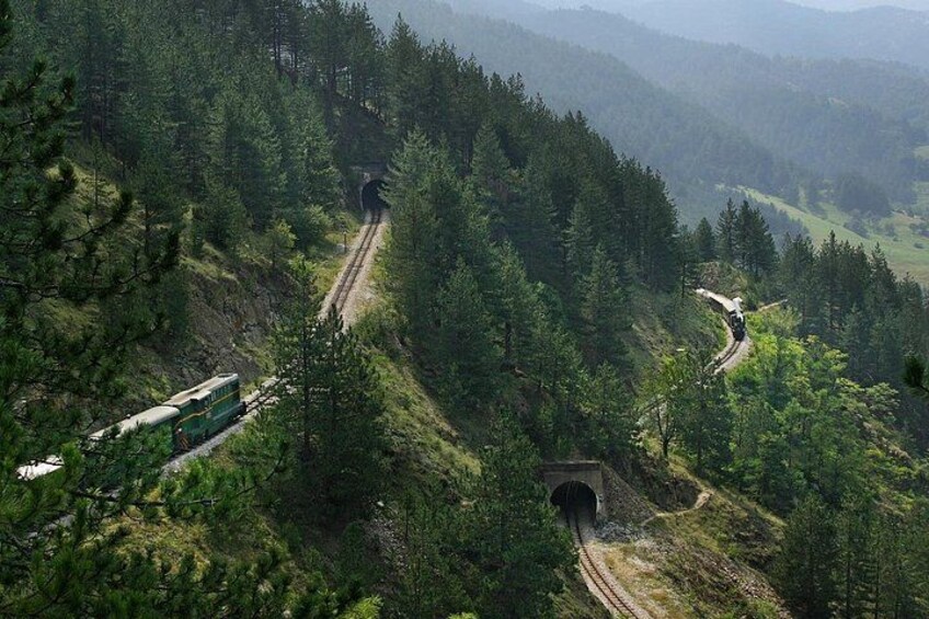 A train traveling through a forested mountainous area with a tunnel and winding tracks. 5