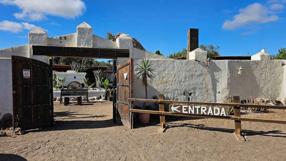 A gate labeled 'ENTRADA' with a rustic wooden fence, a stone wall, and a clear blue sky. 5