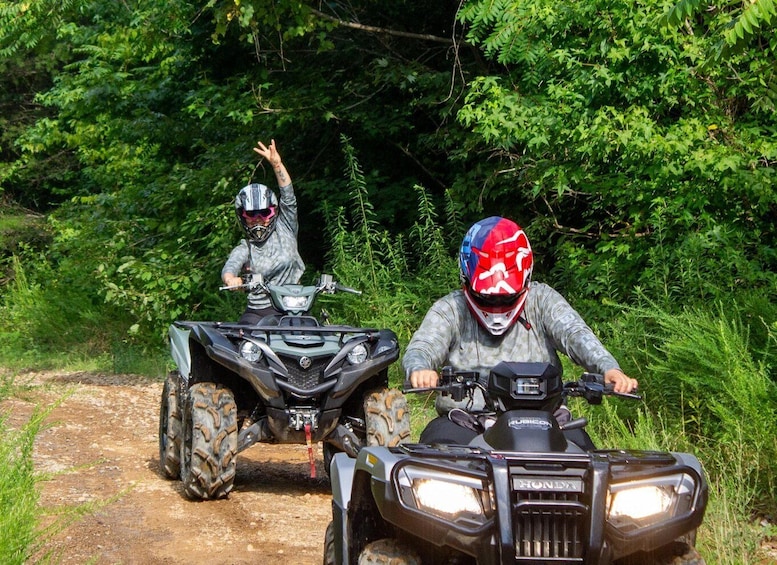 Two people riding ATVs on a dirt path surrounded by dense green foliage. 1