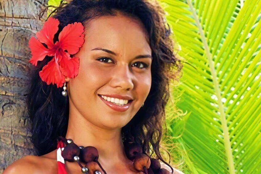 A person with a red flower in their hair, wearing a necklace with beads, and a red and white patterned garment, standing in front of a palm tree. 3
