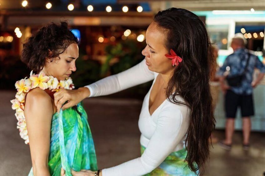 Two people in a public indoor space, one wearing a floral lei and a green dress, the other in a white top. 1