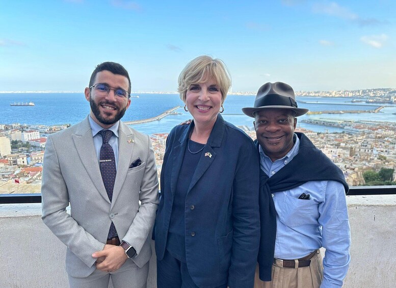 Three people standing on a balcony with a cityscape and port in the background. 4