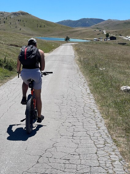 A person riding a bicycle on a paved path with a backpack, surrounded by grassy fields and distant mountains. 1