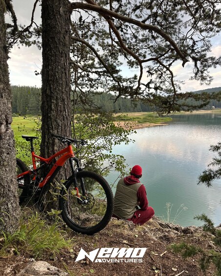 A red mountain bike leaning against a tree by a lake with a person sitting nearby. 5