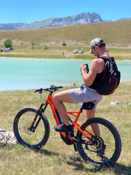 A person sitting on a mountain bike in a grassy field with a lake and mountains in the background. 4