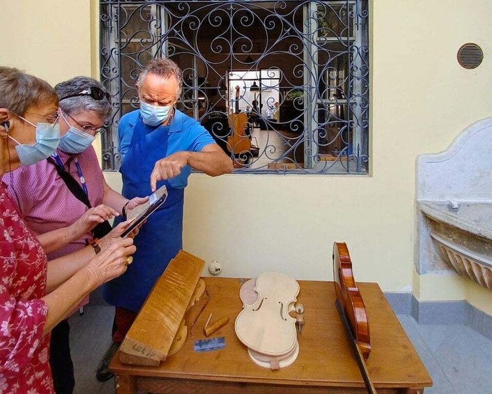 A workshop with a violin on a table, a man pointing at a tablet, and a window with decorative bars. 3