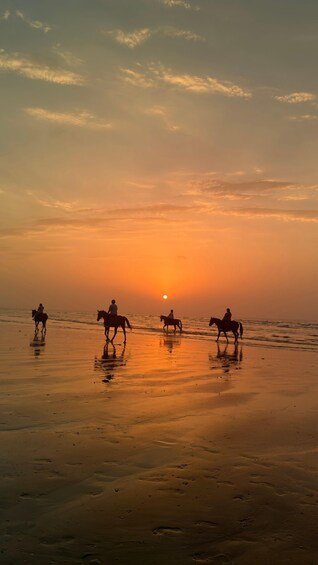 A sunset over a beach with three people riding horses. 1