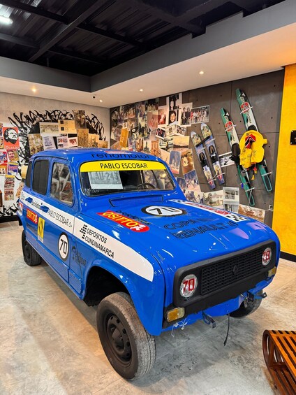 A blue race car with 'Pablo Escobar' and '70' on it, parked indoors with a graffiti-covered wall in the background. 3