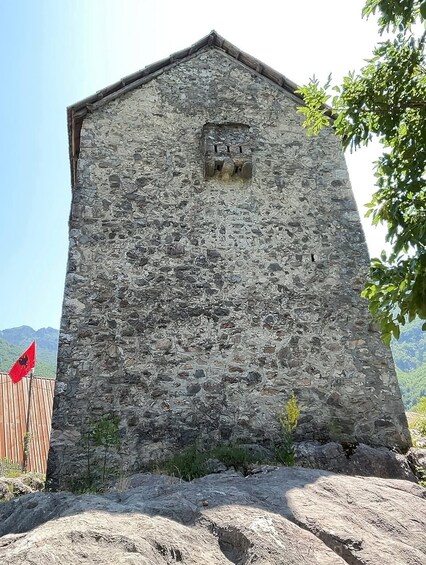 A stone building with a triangular roof, a small window, and a red flag on a wooden post. 5