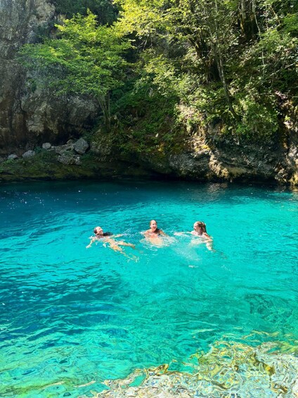 Three people swimming in a clear blue pool surrounded by lush greenery and rocky cliffs. 2