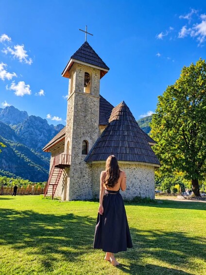 A woman in a black dress standing in front of a church with a bell tower and a cross on top, surrounded by mountains and trees. 1