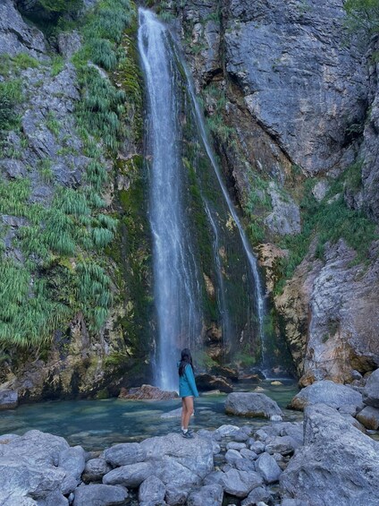 A waterfall cascading down a rocky cliff into a clear pool of water surrounded by large boulders and lush greenery. 3