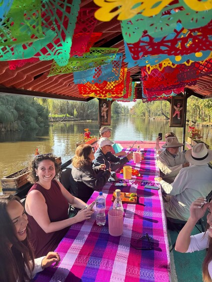 A group of people dining on a boat with a colorful tablecloth, surrounded by a calm river and trees. 2
