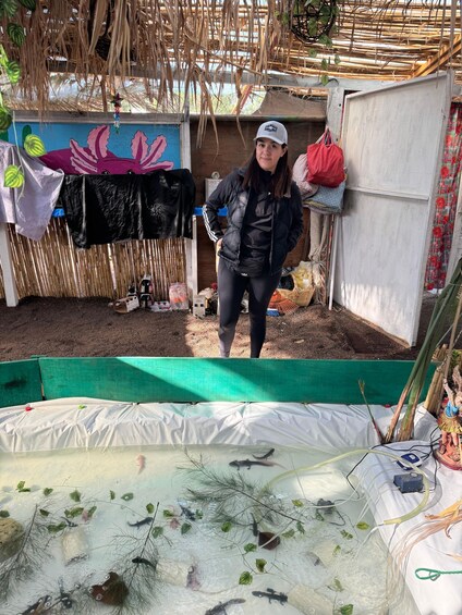 A person standing in a garden with a pond, surrounded by various plants and a bamboo structure. 4