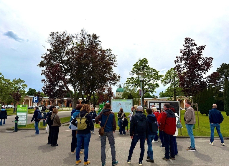 A group of people gathered outdoors, possibly at a public event or meeting, with trees and a building in the background. 2
