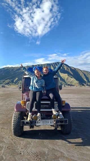 Two people on a vehicle with a license plate, in a parking lot with mountains in the background. 2