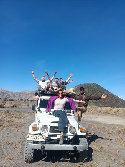 A group of people posing on a white off-road vehicle with a license plate, set against a mountainous landscape. 1