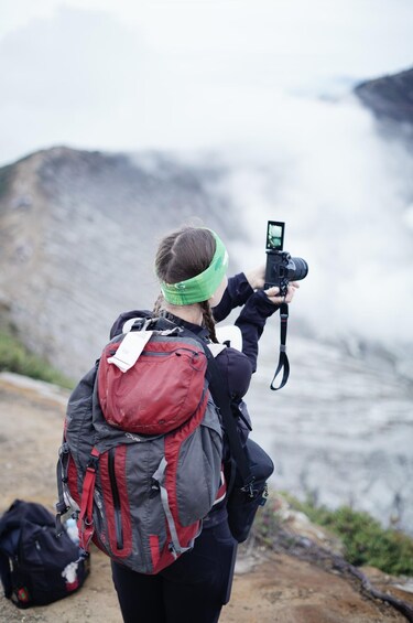 A person with a camera and a green headband, wearing a backpack, taking a photo of a rocky landscape with a body of water. 5