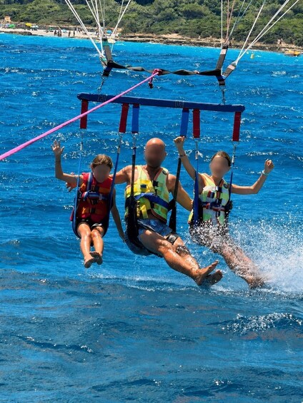 Three people on a water sport activity, wearing life jackets, with a clear blue sea and a rocky coastline in the background. 3
