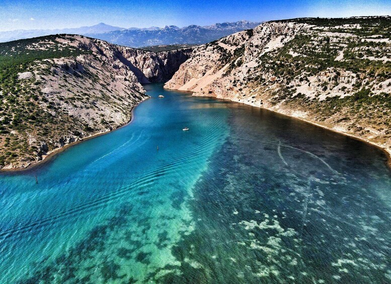 A clear blue sea with a boat, surrounded by rocky terrain and mountains in the background. 1
