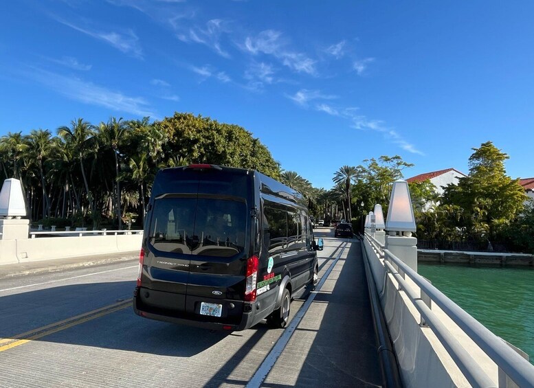 A black van driving on a bridge with palm trees and a building in the background. 1