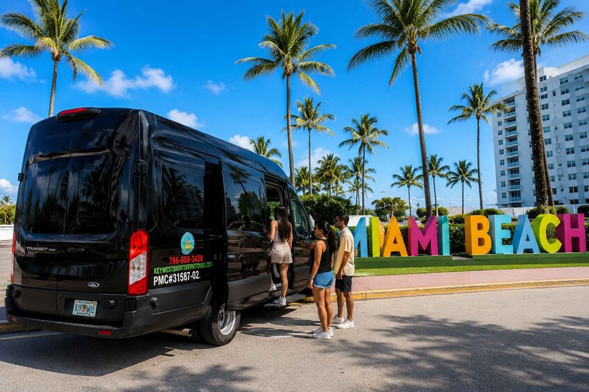 A black van parked in front of a colorful sign that reads 'MIAMI BEACH' with palm trees and a building in the background. 1