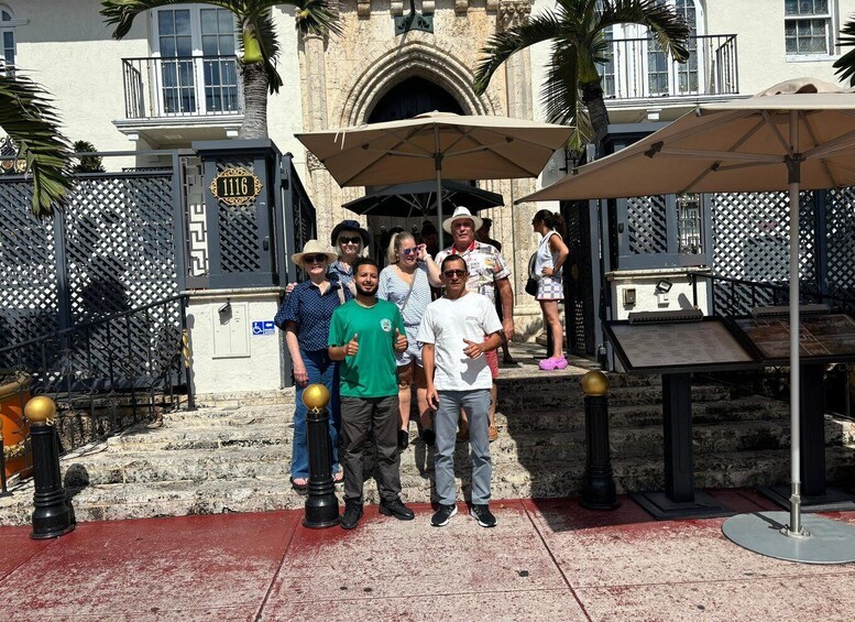 A group of people posing in front of a building with a number sign, a palm tree, and a fence. 2