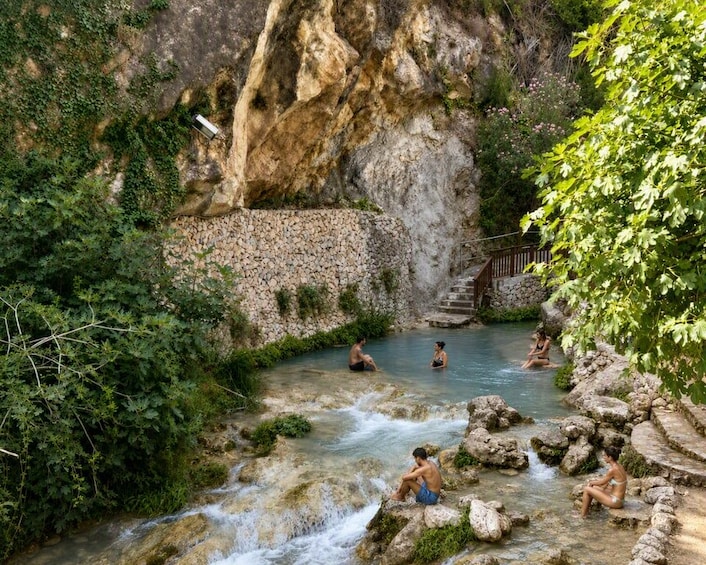A natural hot spring with people relaxing in the water, surrounded by greenery and a stone wall. 2