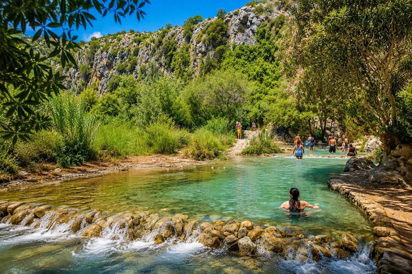 A clear stream with people swimming and walking in a natural setting with rocky cliffs and greenery. 1
