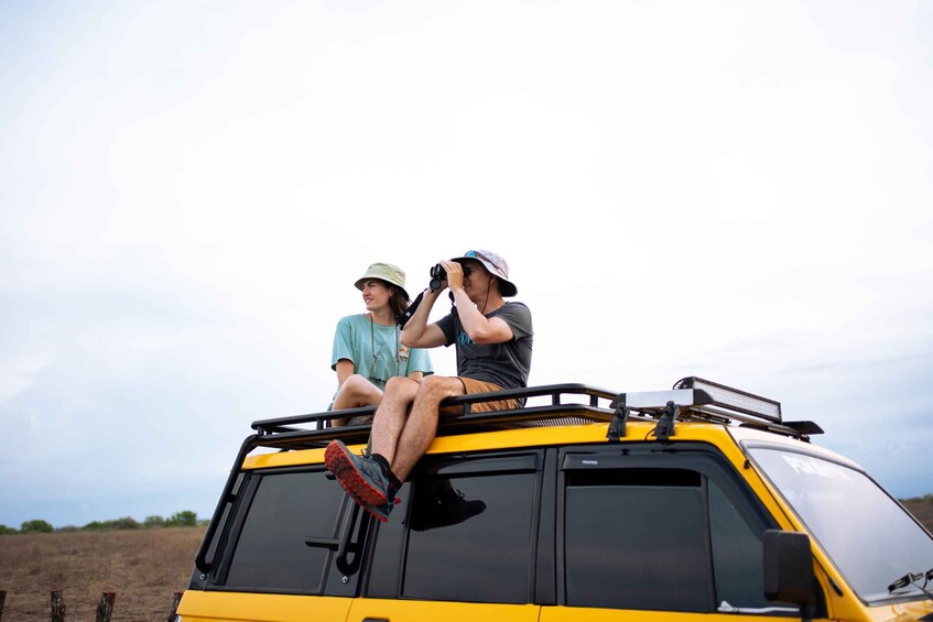 Two people sitting on the roof of a yellow vehicle in an open field. 1