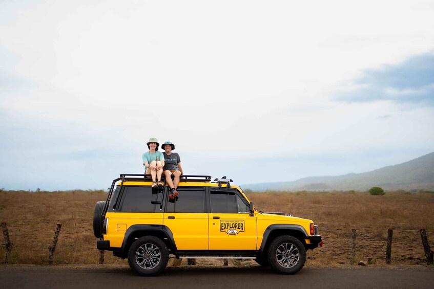 A yellow SUV with two people sitting on the roof, parked on a road with a fence and dry grass field in the background. 4