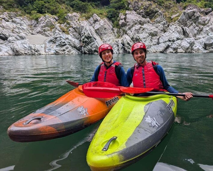 Two people in kayaks on a calm river with rocky banks. 1