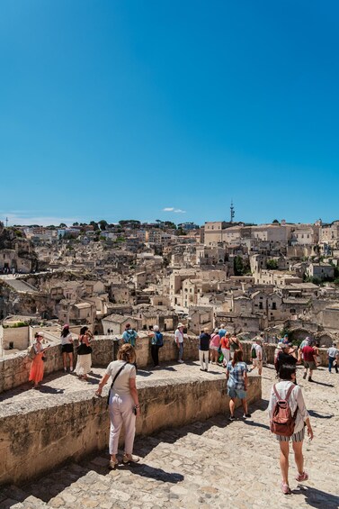A group of tourists walking along a stone pathway overlooking a historic cityscape with ancient buildings and a clear blue sky. 2
