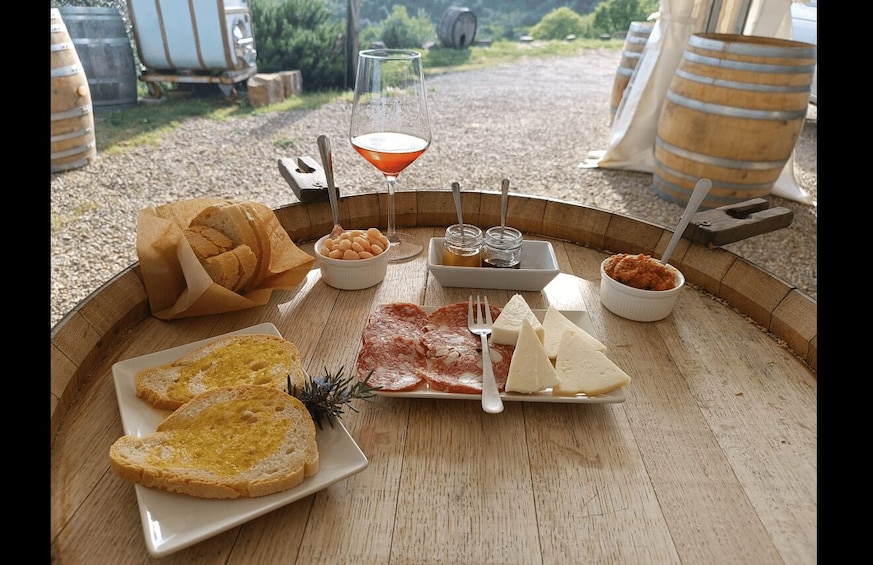 A picnic setup with bread, cheese, cured meats, a glass of red wine, and condiments on a wooden table outdoors. 2