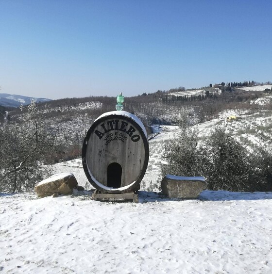 A wooden barrel with 'ALTERO VINE & OIL' written on it, placed on a snowy hill with rocks and trees in the background. 4