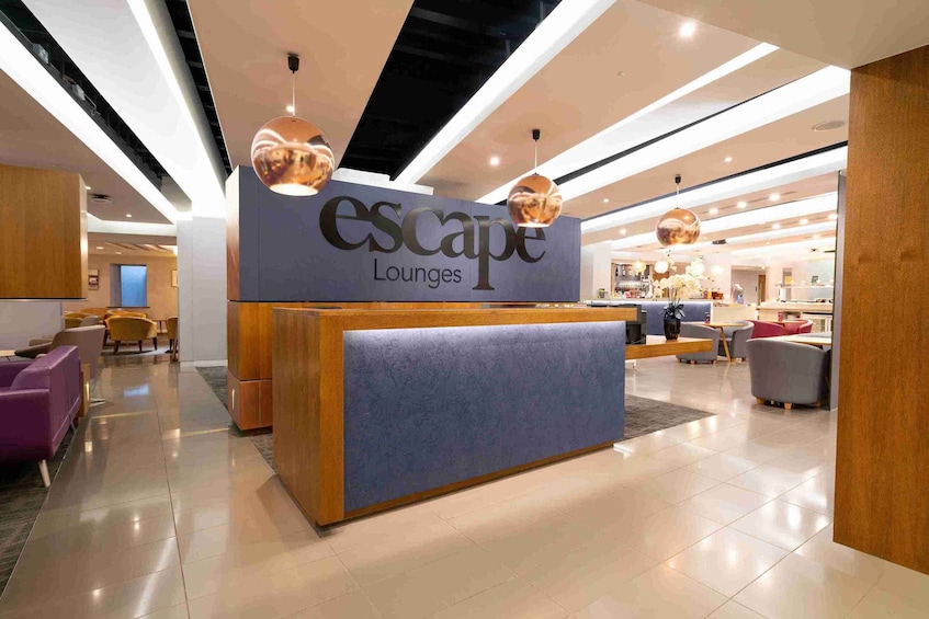 A modern lounge area with a reception desk labeled 'Escape Lounges.' There are multiple seating areas with sofas and chairs, and the space is well-lit with ceiling lights. 1