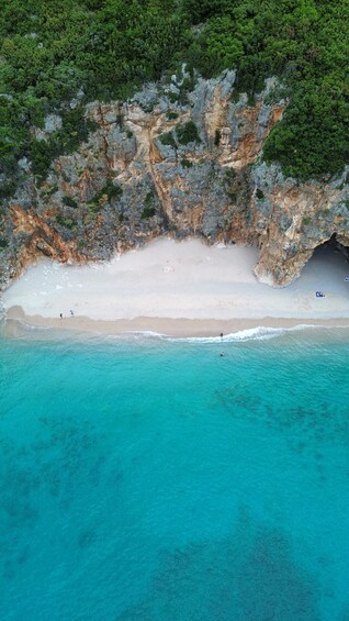 An aerial view of a beach with clear turquoise waters, a rocky cliff with sparse vegetation, and a few people on the shore. 1