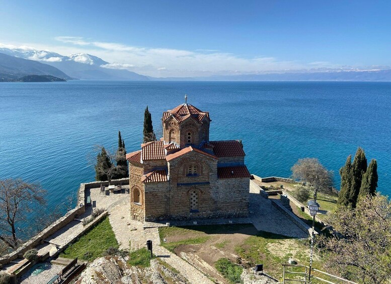 A historic stone church with a red-tiled roof, surrounded by a stone wall and a clear view of a large body of water with mountains in the background. 1