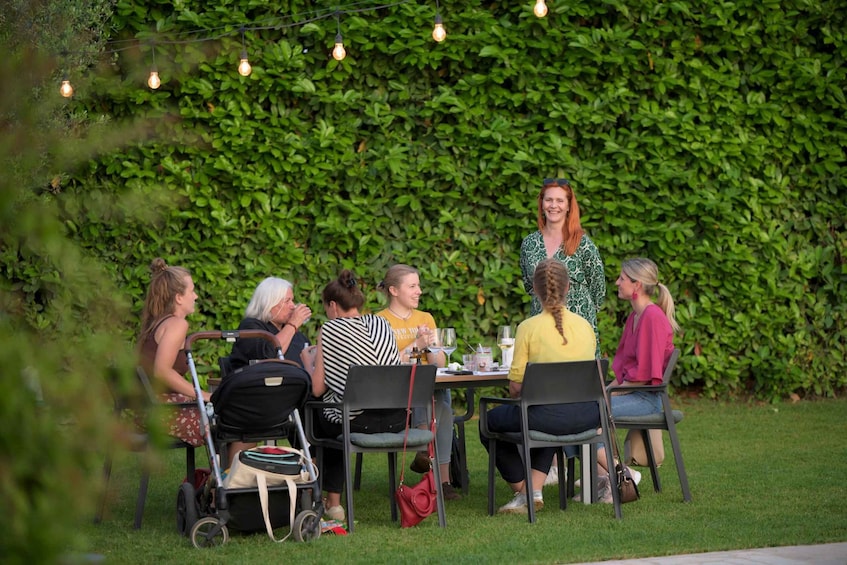 A group of people seated at an outdoor table with a green hedge in the background. 1
