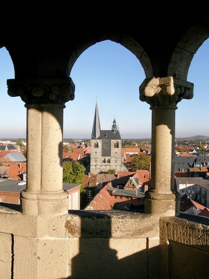 A stone archway framing a view of a church with a spire and clock tower, surrounded by red-roofed buildings. 4