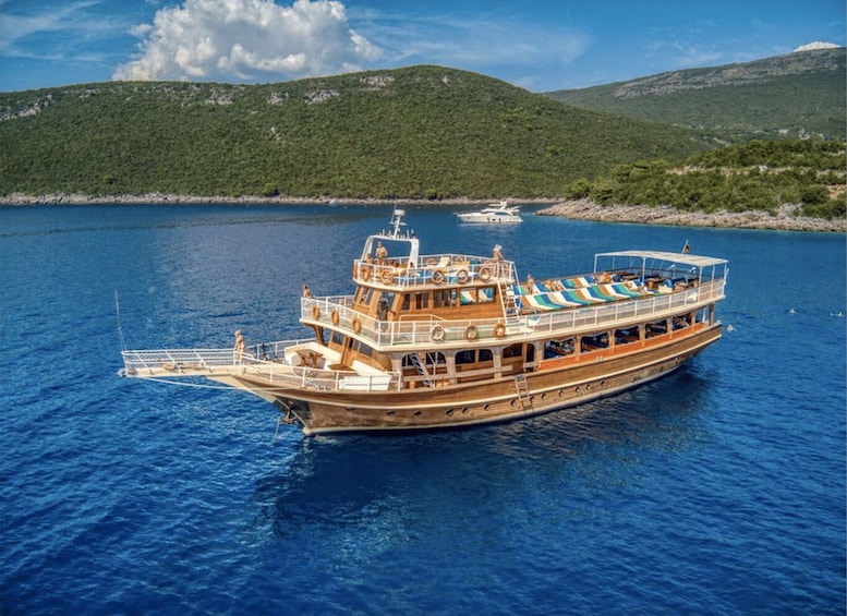 A large wooden boat with multiple decks and a covered upper deck, floating on calm blue waters with a mountainous landscape in the background. 1