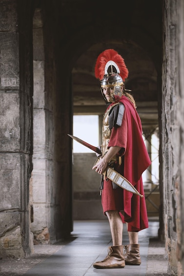 A person in a historical costume with a red cape and helmet, holding a sword, standing in an ancient stone corridor. 2